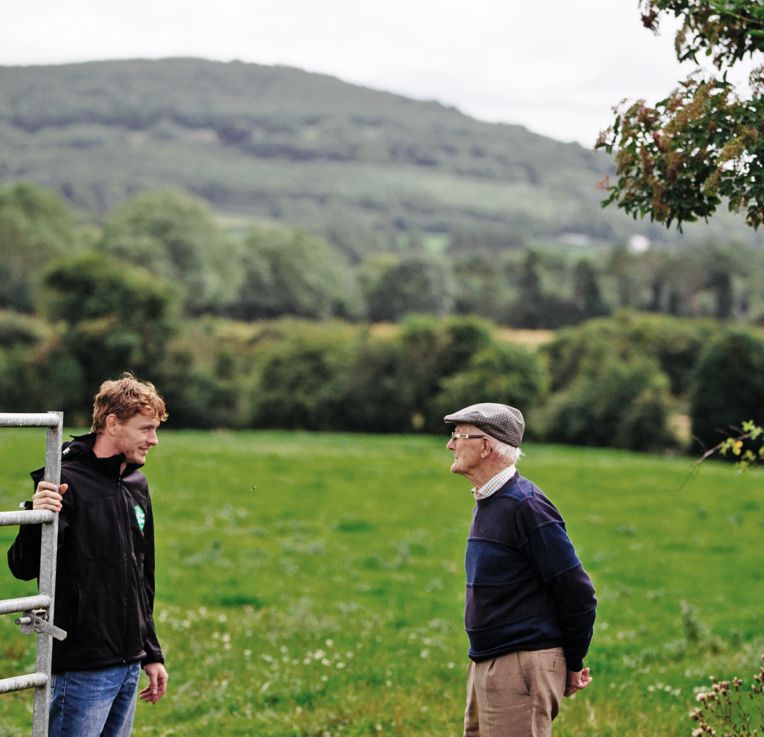 En ung och en äldre jordbrukare samtalar på en grön betesmark i Irland, med kullar i bakgrunden